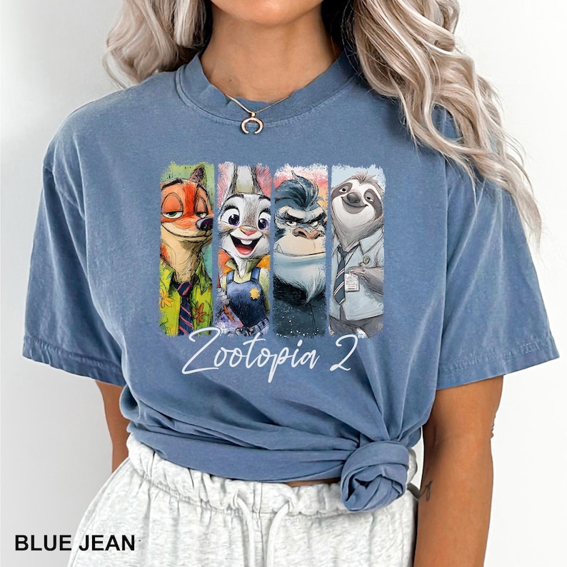Puede incluir: Camiseta azul vaquero con un gr&aacute;fico colorido de personajes de Zootopia 2. El gr&aacute;fico incluye a Nick Wilde, Judy Hopps, un rinoceronte y Flash el perezoso. El texto "Zootopia 2" est&aacute; escrito en cursiva.