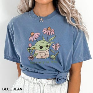Puede incluir: Camiseta azul vaquero con un gr&aacute;fico de un personaje verde con aspecto de extraterrestre rodeado de flores rosas. El texto "ENJOY THE LITTLE THINGS" est&aacute; impreso debajo. La camiseta es de manga corta y cuello redondo.