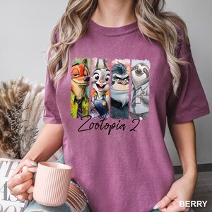 Puede incluir: Camiseta de color bayas con un gr&aacute;fico colorido de personajes de Zootopia 2. El dise&ntilde;o incluye a Nick Wilde, Judy Hopps, Chief Bogo y Flash. El texto "Zootopia 2" est&aacute; impreso debajo de los personajes.
