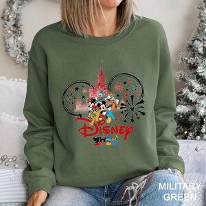 May include: Military green sweatshirt featuring a Disney design. The graphic includes the Disney castle, Mickey Mouse, Donald Duck, and other characters. The word "Disney" is in red, with "2023" below. The design is framed by Mickey Mouse ears and fireworks.