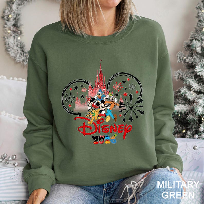 May include: Military green sweatshirt featuring a Disney design. The graphic includes the Disney castle, Mickey Mouse, Donald Duck, and other characters. The word "Disney" is in red, with "2023" below. The design is framed by Mickey Mouse ears and fireworks.