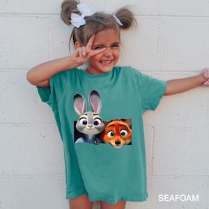 Pu&ograve; includere: T-shirt turchese a maniche corte con Judy Hopps e Nick Wilde di Zootopia. La grafica mostra i personaggi che sbucano da un rettangolo strappato. Il testo "SEAFOAM" &egrave; stampato in basso sulla maglietta.