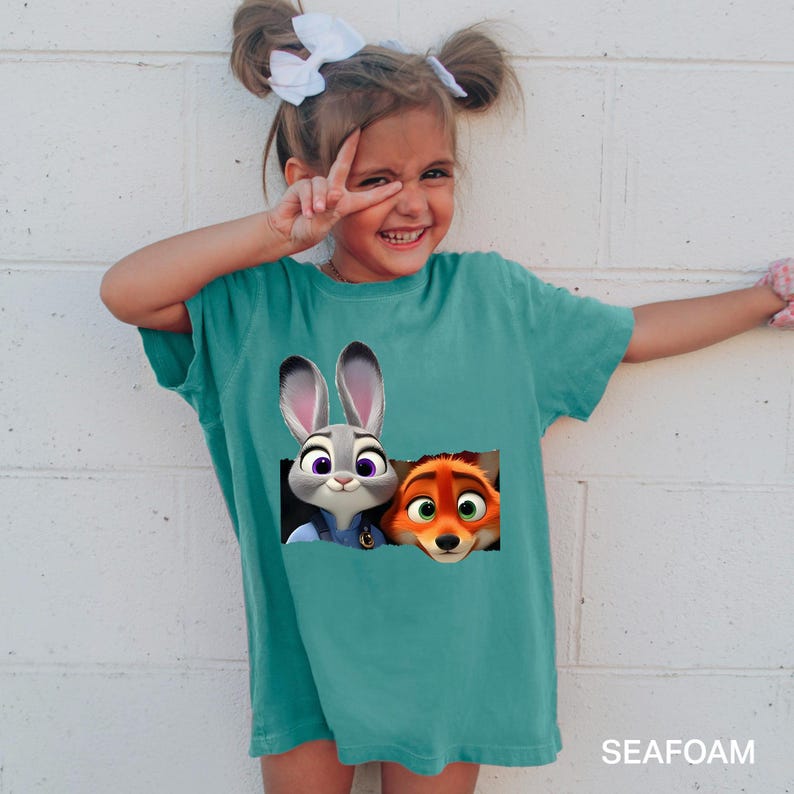 Pu&ograve; includere: T-shirt turchese a maniche corte con Judy Hopps e Nick Wilde di Zootopia. La grafica mostra i personaggi che sbucano da un rettangolo strappato. Il testo "SEAFOAM" &egrave; stampato in basso sulla maglietta.