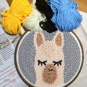May include: A close-up of a llama punch needle kit. The kit includes a wooden embroidery hoop with a gray and beige llama design, four skeins of yarn in yellow, white, blue, and black, and instructions.