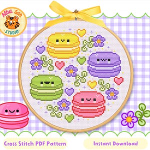Op de afbeelding: Een kruissteek PDF-patroon met een cirkelvormig ontwerp met vier kleurrijke macarons: geel, groen, paars en roze. Het ontwerp bevat harten, bloemen en de tekst "Cross Stitch PDF Pattern" en "Instant Download".