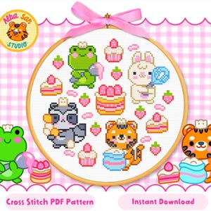 May include: A circular wooden embroidery hoop containing a cross-stitch pattern featuring a green frog, a white rabbit, a grey raccoon, and an orange tiger surrounded by various cakes, cupcakes, and strawberries, topped with a pink ribbon bow.