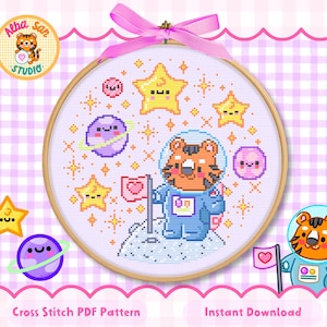Könnte beinhalten: Ein Kreuzstichmuster in einem Holzrahmen mit einem Tiger-Astronauten im blauen Raumanzug auf einer Mondoberfläche mit einer rosa Herzflagge. Das Design zeigt gelbe Sterne, violette Planeten und rosa Kugeln auf einem weißen Gitterhintergrund.