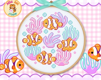 Op de afbeelding: Een ronde houten borduurring met een kruissteekpatroon van vier oranje en witte anemoonvissen die tussen roze en lichtblauw koraal zwemmen. Een lichtblauw lint is bovenaan tot een strik gebonden. De tekst Alba San Studio is zichtbaar.