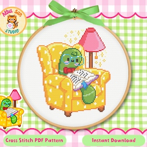 Puede incluir: Un patrón de punto de cruz en un bastidor de madera que muestra a un personaje verde con gafas leyendo un libro en un sillón amarillo con lunares blancos. Una lámpara rosa se sitúa detrás del sillón y una cinta verde está atada arriba.