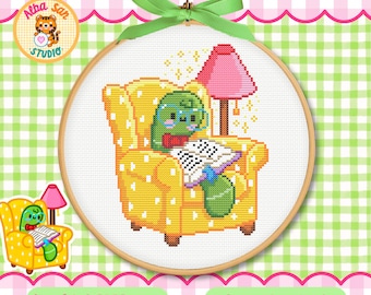 Op de afbeelding: Een kruissteekpatroon in een houten borduurring met een groen figuurtje met een bril dat een boek leest in een gele fauteuil met witte stippen. Een roze lamp staat achter de stoel en een groen lint is aan de bovenkant vastgeknoopt.