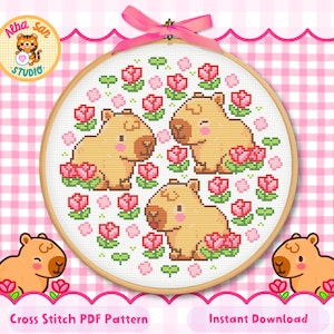 May include: A circular wooden embroidery hoop containing a cross stitch pattern of three light brown capybaras surrounded by pink tulips and small green clover leaves. A pink ribbon is tied in a bow at the top of the hoop. The text Alba San Studio is visible.