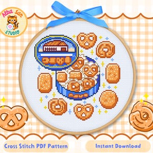 May include: A cross-stitch pattern in a wooden embroidery hoop featuring a blue tin filled with various light brown biscuits and pretzels, topped with a blue ribbon. The design includes scattered small yellow stars and a circular logo with a tiger.