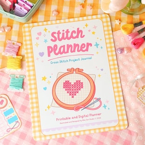 Könnte beinhalten: Ein Stitch Planner-Journal mit einem herzförmigen Kreuzstich-Design. Das Cover zeigt den Text "Stitch Planner" und "Cross Stitch Project Journal". Das Bild enthält Garnrollen, eine Nadel und andere Bastelmaterialien, alles auf einem karierten Hintergrund.