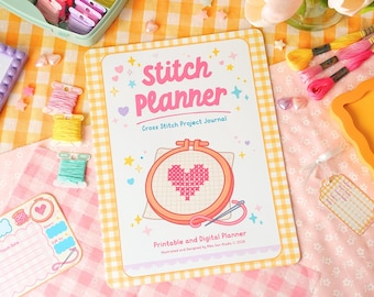 Op de afbeelding: Een Stitch Planner-dagboek met een hartvormig kruissteekontwerp. De omslag bevat de tekst "Stitch Planner" en "Cross Stitch Project Journal". De afbeelding bevat spoelen garen, een naald en andere knutselbenodigdheden, allemaal op een geruite achtergrond.