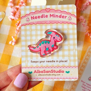 Op de afbeelding: Een roze en blauwgroene dinosaurus naaldhouder op een kaart met de tekst "Needle Minder" en "Keeps your needle in place!". De kaart heeft een ruitpatroon en de winkelnaam "AlbaSanStudio" staat onderaan.