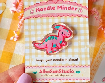 May include: A pink and teal dinosaur needle minder on a card with the text "Needle Minder" and "Keeps your needle in place!". The card has a gingham pattern and the shop name "AlbaSanStudio" is printed on the bottom.