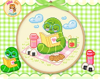Op de afbeelding: Een kruissteekpatroon in een houten borduurring met een groene worm met een blauwe bril die een geel boek met een hart leest. De scène bevat een roze thermoskan, een sapverpakking, twee rijstballen en koekjes op een roze geblokt picknickkleed.