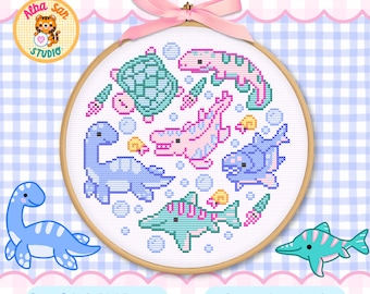 Op de afbeelding: Een ronde houten borduurring met een kruissteekpatroon van diverse aquatische dinosaurussen en zeedieren in pasteltinten blauw, roze en groenblauw, geaccentueerd met kleine schelpjes en bubbels, afgewerkt met een lichtroze strik.