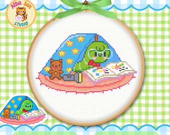 Op de afbeelding: Een kruissteekpatroon in een houten borduurring met een groene rups met een bril die een boek leest onder een blauwe deken met sterren, een bruine teddybeer en een zaklamp op een roze rond kleed, afgewerkt met een groen lint.