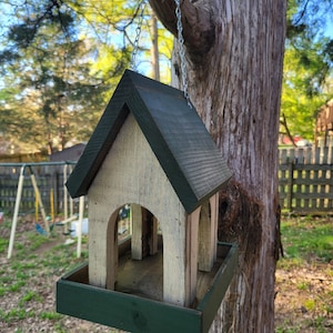 May include: A wooden bird feeder with a green roof and a white base. It has two feeding stations and is hanging from a chain on a tree.