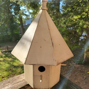 May include: A white wooden birdhouse with a peaked roof and a decorative finial. The birdhouse has a round entrance hole and a small perch.