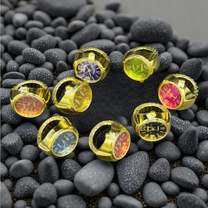 May include: Eight gold ring watches with different colored faces. The rings are on a bed of small gray stones.