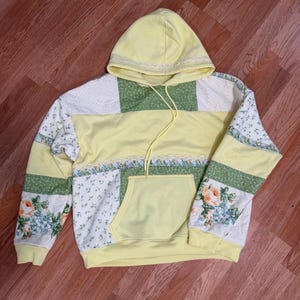 Könnte beinhalten: Ein gelber Patchwork-Hoodie mit einer Vordertasche und Kordelzug. Die Ärmel und die obere Brustpartie sind mit floralen und grünen Stoffen versehen. Die Kapuze ist mit Spitze verziert.