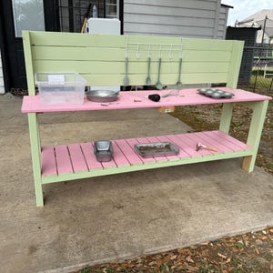 May include: A two-tiered, pink and green wooden play kitchen. The top level has a clear plastic container, a metal bowl, and a muffin tin. The bottom level has a metal container and a small tray. Utensils hang on the back.