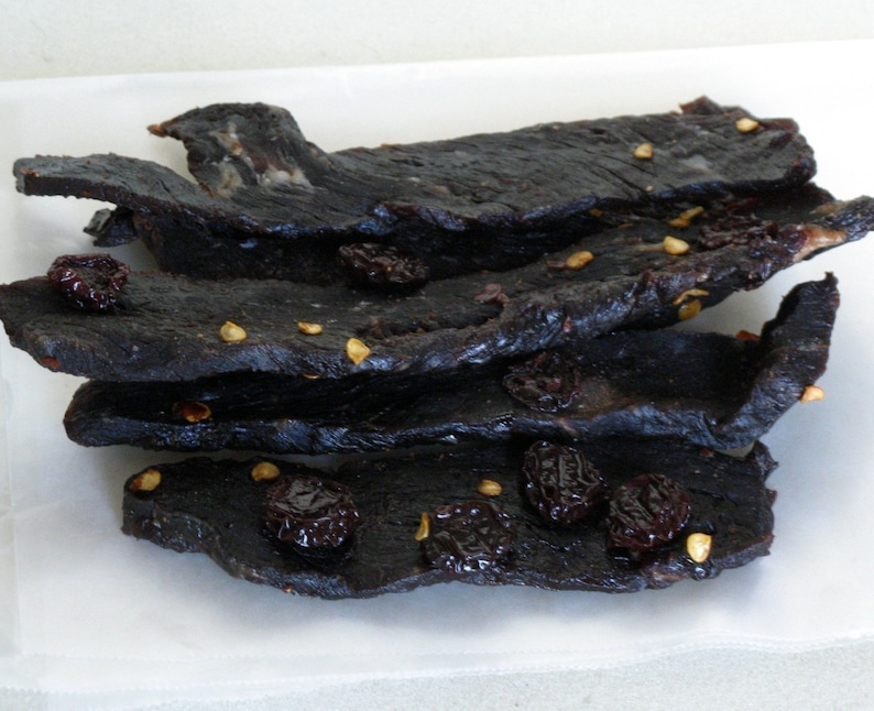 Puede incluir: Una pila de tiras de carne seca oscura con ar&aacute;ndanos secos y granos de pimienta. La carne es de color marr&oacute;n oscuro y brillante, y los ar&aacute;ndanos son de color rojo oscuro.