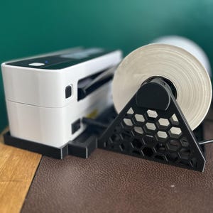 May include: A white label printer with a black base and a roll of white labels. The label roll is held by a black, geometric stand. The printer is on a brown surface.