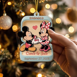 May include: A light blue Christmas ornament featuring Mickey Mouse proposing to Minnie Mouse. The ornament is shaped like a ring box with the word "ENGAGED" at the top. The names "Charlie & Julia" and the date "September 27, 2025" are also displayed.