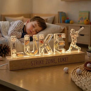 May include: A wooden baseball-themed night light spelling out the name "LUKE" with baseballs, bats, and a baseball player silhouette. The base reads "STRIKE ZONE, LUKE!" A sleeping child is in the background.