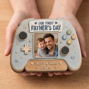 May include: A wooden Father's Day photo frame shaped like a video game controller. The frame features a photo of a father and child, with the text "OUR FIRST FATHER'S DAY" and "DADDY & NOAH 2026". The controller is light blue and wood-toned.