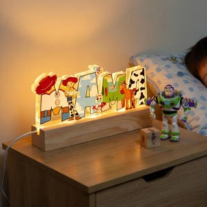 May include: A wooden night light with the name "AMA" illuminated, featuring characters from the Toy Story movie. A sleeping child cuddles a Woody doll. A Buzz Lightyear figure and a Toy Story poster are also visible.