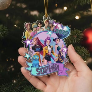 May include: A clear acrylic Christmas ornament featuring a group of animated characters. The ornament has a purple and teal banner with the name "SOPHIA". A disco ball with "2025" is also featured. The ornament is held in front of a Christmas tree.