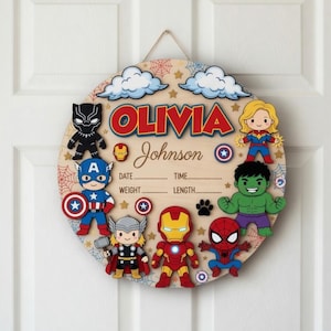 May include: A round wooden sign with the name "Olivia Johnson" in red letters, surrounded by cartoon superhero characters. Includes spaces to record birth details. The sign is decorated with clouds, stars, and spiderwebs.