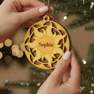 May include: A personalized wooden Christmas ornament with a sun design. The ornament is yellow and brown, with the name "Sophia" in the center. It is held by a person in front of a Christmas tree.