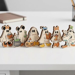 Pode incluir: Um conjunto de seis figuras de madeira recortadas com personagens de desenhos animados vestidos de fantasmas para o Halloween. Cada figura exibe detalhes como abóboras, caldeirões e baldes de doces, com o nome James impresso em várias peças.