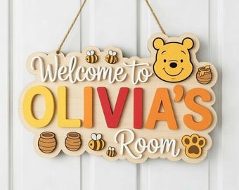 Może przedstawiać: Drewniany znak z napisem "Welcome to OLIVIA'S Room" w kolorze białym, żółtym, pomarańczowym i czerwonym. Zawiera twarz misia z kreskówki, słoik miodu, pszczoły i odcisk łapy. Znak jest zawieszony na naturalnym sznurku.