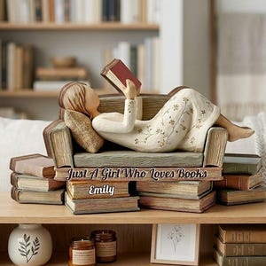 Puede incluir: Figura decorativa de una persona leyendo un libro, reclinada en un sofá hecho de libros. La figura lleva un vestido blanco con motivos florales. El texto "Just A Girl Who Loves Books" y "Emily" son visibles.
