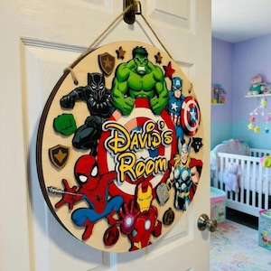 May include: A circular wooden sign decorated with Marvel superheroes such as Spider-Man and Iron Man. The sign displays the text "David's Room" in a colourful, cartoon style. It is suspended from a white door by a rope.
