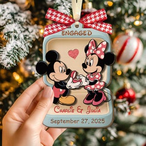 May include: Wooden Christmas ornament featuring Mickey Mouse proposing to Minnie Mouse. The ornament has a red and white bow, the word "ENGAGED", a heart, and the names "Charlie & Julia" with the date "September 27, 2025".