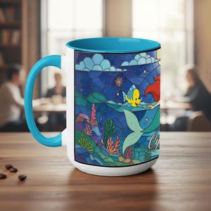 Puede incluir: Taza de cer&aacute;mica blanca con interior y asa azules. La taza presenta una colorida ilustraci&oacute;n de una sirena, un pez amarillo y un paisaje submarino. El dise&ntilde;o recuerda a las vidrieras, con un fondo de cielo estrellado.