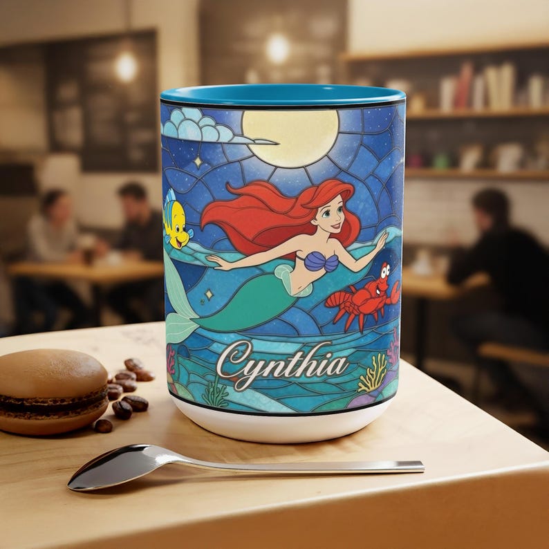 Puede incluir: Taza de caf&eacute; cil&iacute;ndrica con una ilustraci&oacute;n de estilo vidriera de Ariel de La Sirenita. El dise&ntilde;o incluye a Ariel, Flounder, Sebasti&aacute;n y el nombre "Cynthia". La taza tiene un interior azul y una base blanca.