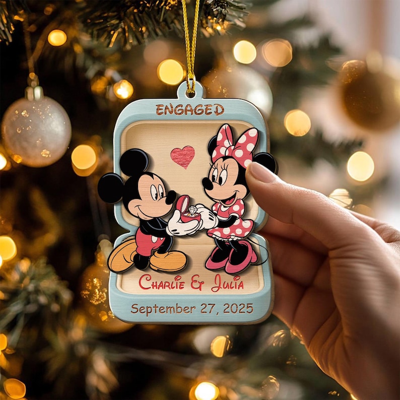 May include: A light blue Christmas ornament featuring Mickey Mouse proposing to Minnie Mouse. The ornament is shaped like a ring box with the word "ENGAGED" at the top. The names "Charlie & Julia" and the date "September 27, 2025" are also displayed.