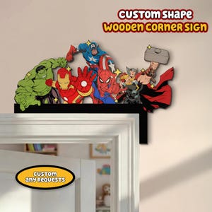 Könnte beinhalten: Ein individuell geformtes Eckschild aus Holz mit einem farbenfrohen Design von mehreren Superhelden, darunter Hulk, Iron Man, Captain America und Spider-Man. Das Schild ist schwarz und an einer Wand über einer Tür angebracht. Das Schild trägt den Text "CUSTOM SHAPE WOODEN CORNER SIGN".