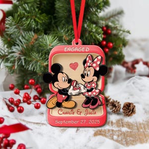 May include: A red and wooden Christmas ornament featuring Mickey Mouse proposing to Minnie Mouse. The ornament is shaped like a jewelry box with the word "ENGAGED" at the top. The names "Charlie & Julia" and the date "September 27, 2025" are also displayed.