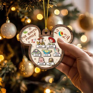 Puede incluir: Un adorno navideño de madera con forma de cabeza de Mickey Mouse. El adorno está decorado con una familia de personajes de dibujos animados de la película Toy Story. El adorno está personalizado con el texto "The Smith Family" y el año "2024".