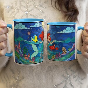 Puede incluir: Dos tazas de cer&aacute;mica con interior y asa azules, con un dise&ntilde;o de estilo vidriera. Una taza representa una sirena con un pez y la otra una sirena con un cangrejo. Las tazas tienen un borde y un asa azules.