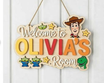 May include: Wooden sign with the text "Welcome to OLIVIA'S Room" in a playful font. Features cartoon characters, including Woody, aliens, and a dinosaur. The sign is designed with a light wood base and colorful accents.